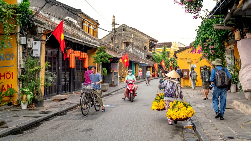 Strada nella città vecchia di Hoi An con case gialle, lanterne rosse e bandiere vietnamite, e un venditore ambulante che porta fiori gialli